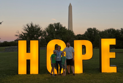family of four, mom, dad son, daughter, in front of a light up sign that says hope